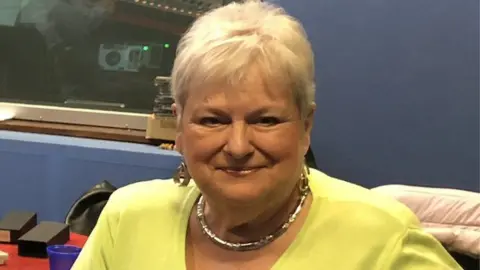 Sally Beardsworth with short blond hair, wearing a yellow top, necklace and large earrings