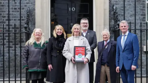 Ben Barnett Three women and three men stand in front of the black Downing Street door. The woman in the centre holds a framed petition.