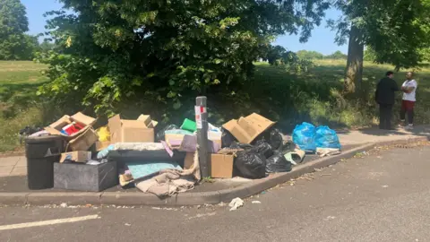 Piles of rubbish on a street in Birmingham. Cardboard boxes, bin bags and other items are piled up on a pavement next to the road. There are trees and a large expanse of grass to the left of the pavement. Two men stand near the rubbish, talking to each other.