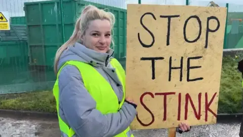 A young woman with long blonde hair, in a grey coat and high vis vest holds a sign that says 'stop the stink' in front of large green skips at the landfill site