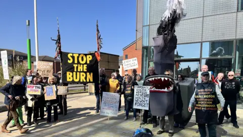 A group of protesters on the pavement outside a building on a sunny day. They are holding signs that say 'stop the burn' and 'clean air for good'. One is dressed like a shark. 