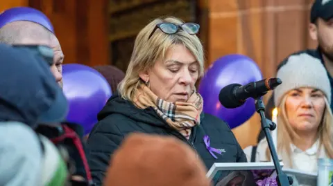 PA Media A woman, wearing a coat and scarf and with glasses on her head, speaks into a microphone. she is surrounded by other people, a number of purple ballons can be seen behind her