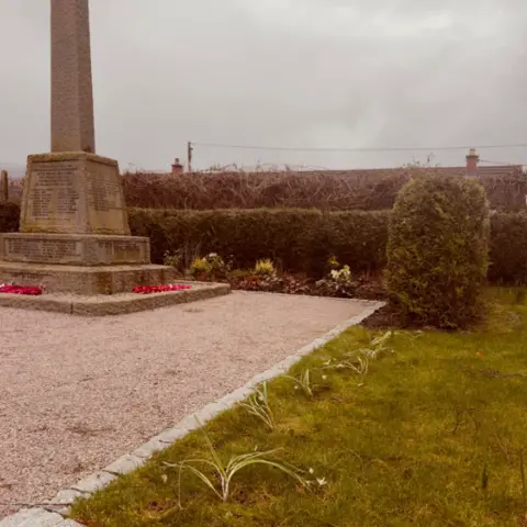 Corrie Snowdrops A war memorial with a gravel path. Beside it is a grassy area where snowdrops have ben planted