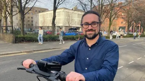 Claire Hamilton/BBC News Richard MacDonald seated on an e-scooter with a dark blue patterned shirt. Richard has dark hair, black spectacles and a beard.