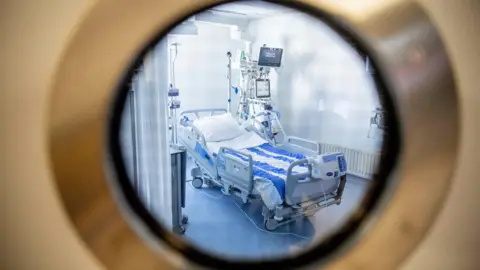 PA Media Looking through a round window into a hospital room which has a bed with wheels with a row of monitors next to it.