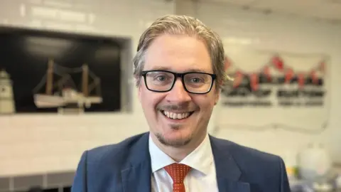Steven Booth has greying hair and smiles for the camera. He is wearing a dark blue suit jacket, a white shirt and a red patterned tie.