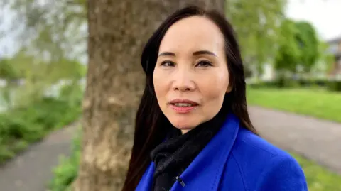 BBC Una mujer con un abrigo azul se para frente a un árbol en un parque mirando a la cámara.