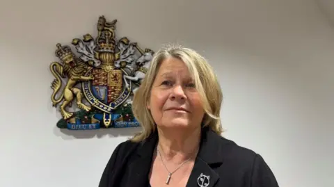 Coroner Johanna Thompson, wearing a black jacket, with silver brooch, stands in the coroner's court by the Royal Crest. She has blonde shoulder length hair, and is wearing a silver pendant necklace.