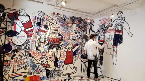 Team GB A man in a white shirt and black trousers. He is painting an expressionist-style mural on a wall.