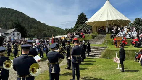 A cream-coloured canopy over the top of Tynwald Hill, which has dignitaries seating on chairs around it and in each tier. Members of the military band, dressed in blue, are standing at the foot of the hill.