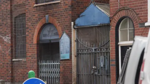 PA Media A close-up of a red brick building featuring an arched entrance with a Star of David and the years 1923 and 5684 inscribed above the door. A blue sign for the "East London Central Synagogue" is mounted on the wall. To the right of the entrance is a tall, padlocked metal gate topped with a weathered blue gabled roof.