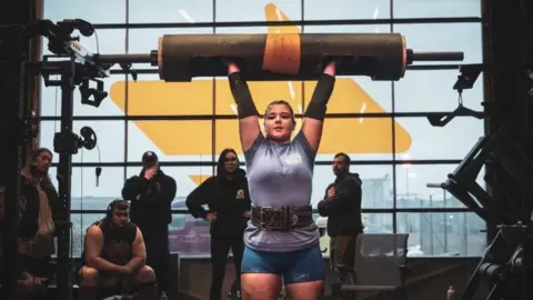 Gordon Siegal Lauri stands in a gym, lifting a large cylindrical bar overhead with both arms extended. Several onlookers and gym equipment surround the platform, with a large window behind letting in muted daylight. There are belts, racks, and machines visible.