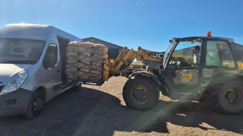 Anna Foster/Whittlesey Community Pantry A yellow farm vehicle with a fork lift on the right loading bags of potatoes into a white van parked on the left. 