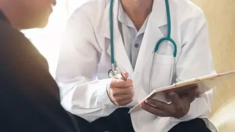 Getty Images A doctor wearing a white coat and a blue stethoscope, holding a clipboard and pen, is talking to a male patient wearing black