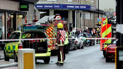 Getty Images A picture from the 7th of July 2005 of the Edgeware Road station after the bombings. it is a large building with a dark blue sign displaying its name in white writing. The area in front of it is cordoned off with red and white scene tape. There are two fire engines, several cars and an ambulane parked with people standing on scene.