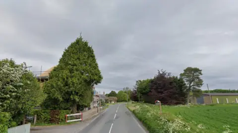 A streetview image of the the A4361 in Winterbourne Monkton. The two-lane road runs past a small village. There is a green field on the right and trees and hedgerows on both sides of the pictures, with some houses and farm buildings. The sky is overcast.  