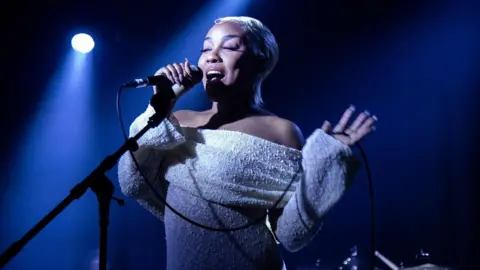 Rosemary Nwodoh is seen performing on a stage a light-coloured off-the-shoulder dress. The stage is lit up in blue and white and she is standing in front of a microphone as she sings.