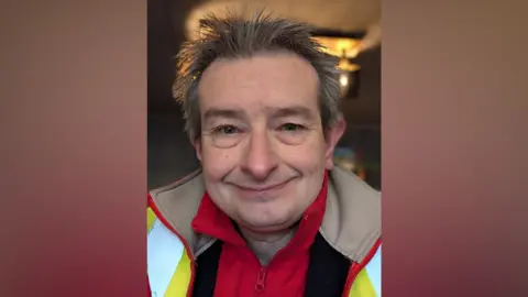 Rich Todd A man with dark grey hair smiles in a selfie. He is wearing a yellow hi-vis jacket with a red fleece underneath.
