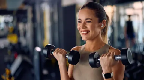 A woman in a gym, who is holding two dumbells and smiling towards the camera. She has a blonde ponytail and is wearing gym clothes.