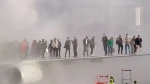 BBC People stand on the wing of a plane on the tarmac. Smoke can be seen encircling around them, whilst airport staff on the ground begin to bring ladders to help them dismount.