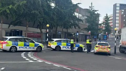 A cordoned off street with police cars and  tape sectioning off a road. There are closed shops on either side of the road in the background. There are also some trees on the left side pavement in the distance. 