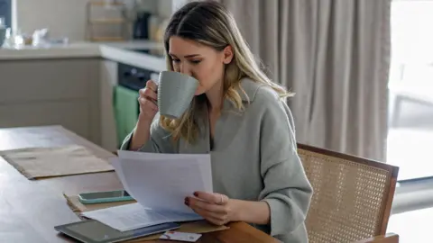 Getty Images Una mujer sentada en una silla en casa mirando papeles y bebiendo una taza de té.