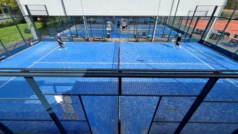 Padel court from above. The court is blue with white lines. It is inside a black fence. People are on the court playing. There is another court in the background with a roof. 