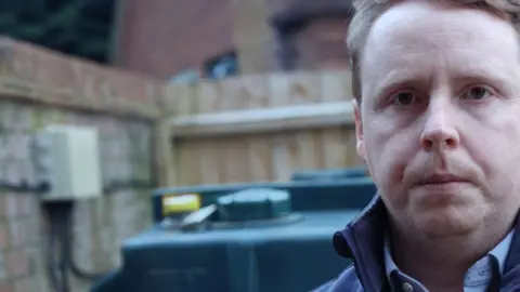 John Birchall John Birchall standing in front of his dark green oil tank. He has light brown hair and is clean shaven. He is wearing a dark blue gilet over a light blue shirt. 