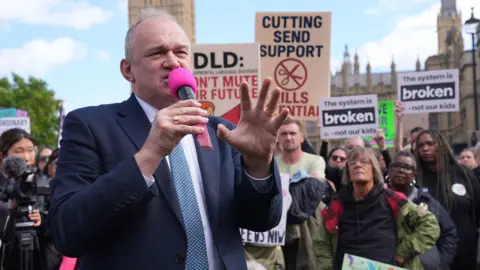 Lib Dem leader Sir Ed Davey speaks into a pink microphone at a rally of SEND campaigners outside the Houses of Parliament. Behind him people hold placards reading "the system is broken - not our kids".