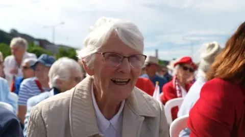Betty Hambly. She has short white hair and glasses. She is smiling and wearing a cream coloured jacket and glasses. 