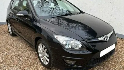 A black Hyundai i30 car with the registration plate blurred out is parked on light-coloured gravel.