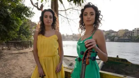 The Ayoub sisters standing by a river, wearing dresses - one yellow, one green, and holding a violin and cello,