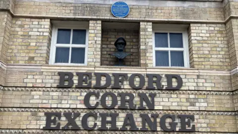 Alex Pope/BBC A blue plaque and statue on the front of Bedford Corn Exchange. The statue is of his head, set back into the wall, which is made of light brick. There are two white windows and signage for the building. 