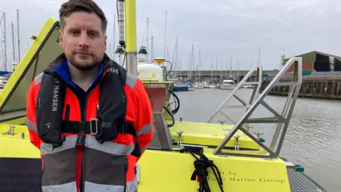 A man in an orange high-vis jacket with a life vest stands with his hands behind his back in front of the yellow vessel. He has short brown hair and a beard and looks seriously at the camera. 
