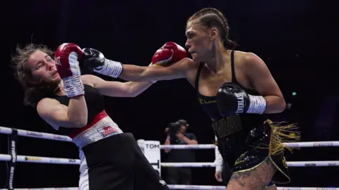 Matchroom Boxing Tiah throwing a punch towards her opponent in the boxing ring. She is wearing black shorts with gold fringing details and black boxing gloves, with her hair in tight plaits.