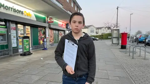 A young boy holding a clipboard and paper outside a Morrisons Daily store 