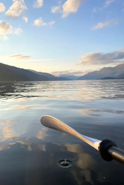 Kathryn Boag The oar of a kayak above still water, with one small drip making a splash, with hills and clouds in the distance.