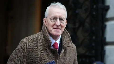 PA Media Hilary Benn has light grey hair and metal-framed glasses and is wearing a brown coat over a blue shirt and red tie