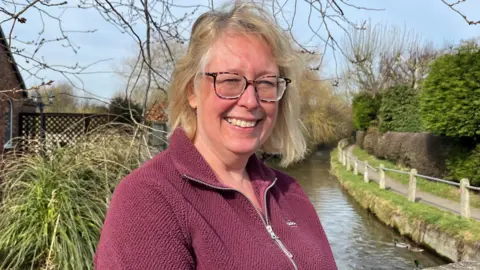 A head and shoulder picture of a middle aged woman with a blond bob, glasses and a dark red fleece top standing on a bridge overlooking a shallow clear stream. There is a fenced pathway by the side of the river to one side of her and a garden filled with plants on the other.