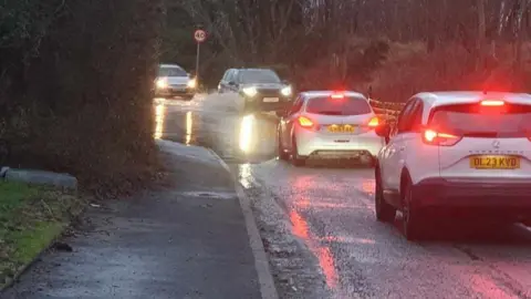 David Eccleston Two cars on the left hand side of Gobowen Road with their red brake lights on as they drive through standing water, which stretches across the width of the road. There are two cars coming in the opposite direction on the right hand side of the road. They have their headlights on. The front car is driving through the water, and there is water splashing up around the front wheel.