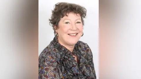 Monmouthshire County Council Sue Riley - a middle aged white woman with short, brown, curly hair. She is posing for a staged photo in front of an all-white background and is wearing a predominantly brown multi-coloured shirt. She has large hooped earrings on and is smiling