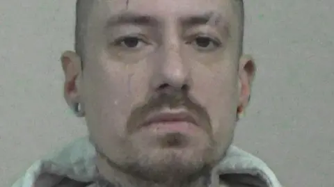 Mugshot of Phillips. He has a short beard and shaved head, with tattoos on his neck and large round earrings in the lobes of his ears