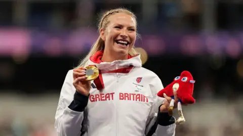 Wheelchair racer Samantha Kinghorn looks ecstatic as she holds up her newly-presented gold Olympic medal on the podium in Paris. She wears her Team GM tracksuit and beams at the appreciative crowd.