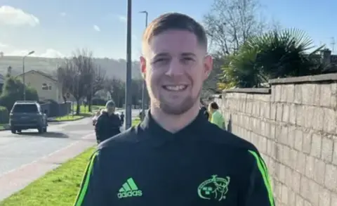 The late Ian Baitson smiling at the camera.  He has short, blond hair and is wearing a black sports top with bright green stripes and logos.  He is standing at the side of a road on a sunny day with a group of people behind him. 