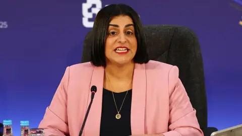 PA Media Home Secretary Shabana Mahmood seated at a conference table, wearing a pink blazer over a dark top and a pendant necklace. A microphone is positioned in front of her. The background is a blue wall featuring a white geometric logo.