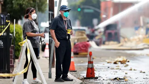Two residents in masks look on as firefighters work on teh fire