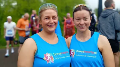 Two women stand side by side wearing matching blue sleeveless running tops with the text “braveheartsni – Run like the wind for Braveheart NI” printed on the front. Both wear sunglasses pushed up on their heads. Other runners in athletic clothing are visible in the background on a tree-lined path, some wearing race bibs and preparing for or taking part in the event.
