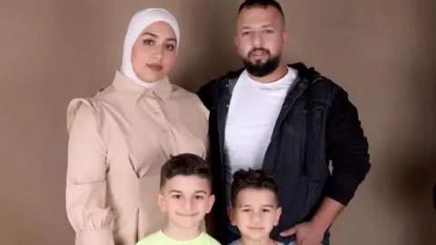 Mahasen Al-Dada  Mahasen al-Dada poses for a photograph in a beige coat, with her arm around her husband. Their two boys stand in front of them.