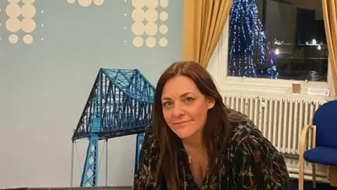 Middlesbrough Council Joanne Rush has a long brown hair and wears a black top with flowers, possibly red roses or tulips, on it.. Behind her is a picture of the Transporter Bridge. A Christmas tree is lit up outside.