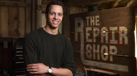 BBC/Ricochet Ltd Will Kirk, with short dark hair and wearing a brown sweater, stands in front of the Repair Shop sign